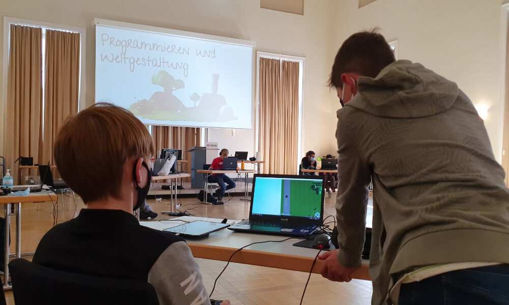 Kinder stehen an einem Tisch mit Laptop und diskutieren über den Inhalt, Auf einer Leinwand im Hintergrund steht "Programmieren und Weltgestaltung"