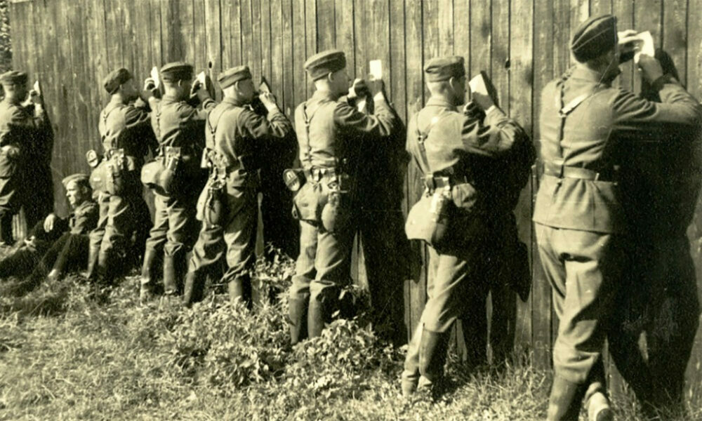 Eine Reihe von Soldaten aus dem 2. Weltkrieg sind von hinten zu sehen. Sie stehen an einer Holzwand aus Latten und schreiben Postkarten. Die Holzwand dient als Schreibunterfläche.