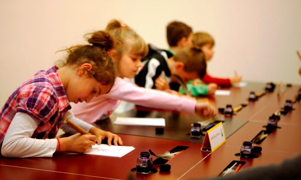 Kinder in der Ausstellung sitzen an Tischen der Schreibwerkschaft und schreiben mit unterschiedlichen Stiften. Museum für Kommunikation