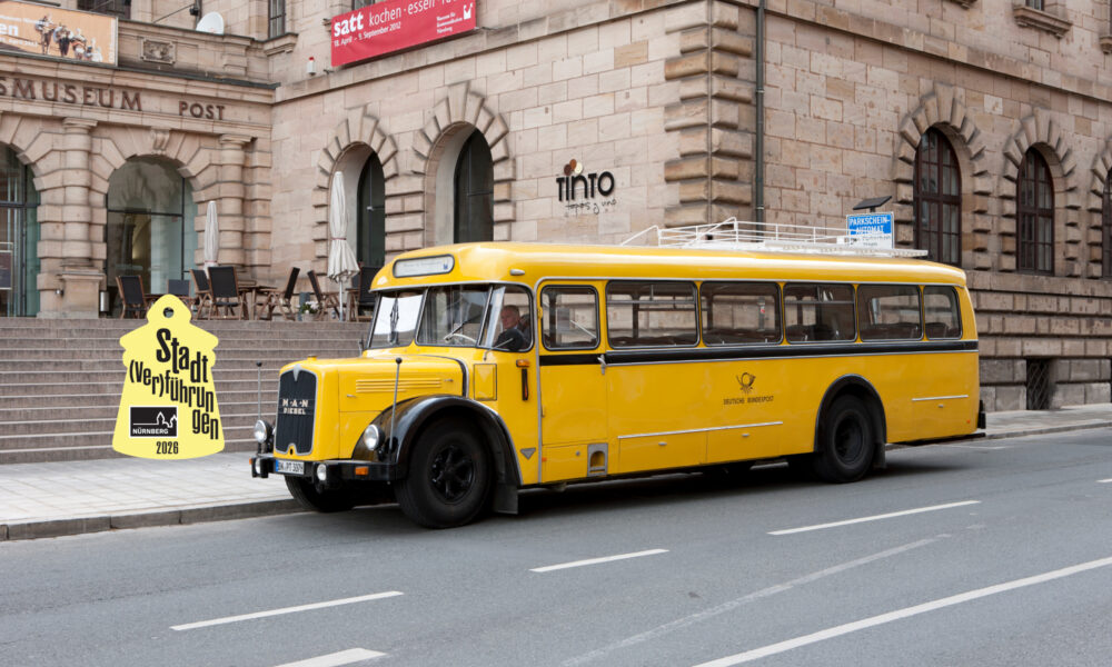 Der MAN Kraftpostbus von 1956 steht vor dem Verkehrsmuseum. Das Emblem von den Stadtverführungen 2026 schwebt über dem Bus