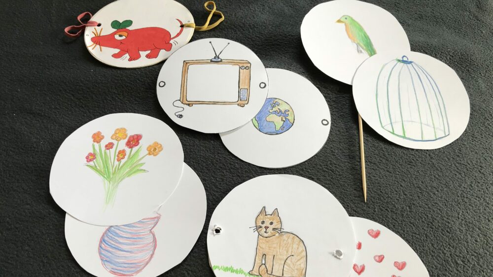 Mit Symbolen und Tieren bemalte Papierkreise, Gummis und ein Holzspieß, die zum erstellen von Wunderscheiben bereit liegen.