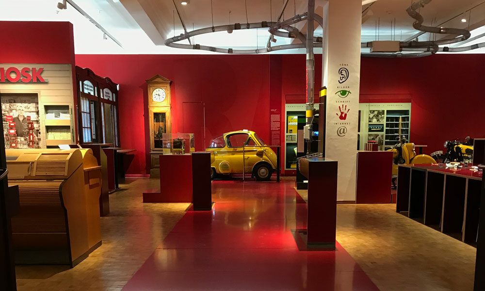 Innenansicht des Museum für Kommunikation, Blick in den roten Bereich der Ausstellung, im Zentrum die kleine gelbe Post-Isetta, links eine historische Post-Theke, am Pfeiler in der Mitte endet die Rohrpost.