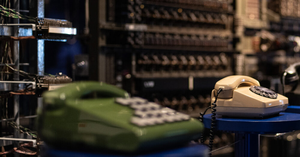 Blick in die Ausstellung im Museum für Kommunikation: grünes Tastentelefon, beiges Telefon mit Wählscheibe, altes Schwarzes Telefon: alle drei Telefone stehe vor einer historischen Vermittlungsanlage