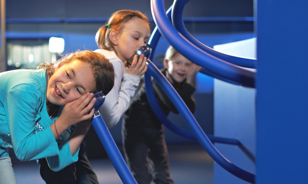 Die Rohrpost im Museum für Kommunikation, Kinder halten ihren Mund oder ihr Ohr an die Trichter, die aus der blauen Ausstellungswand herausragen.